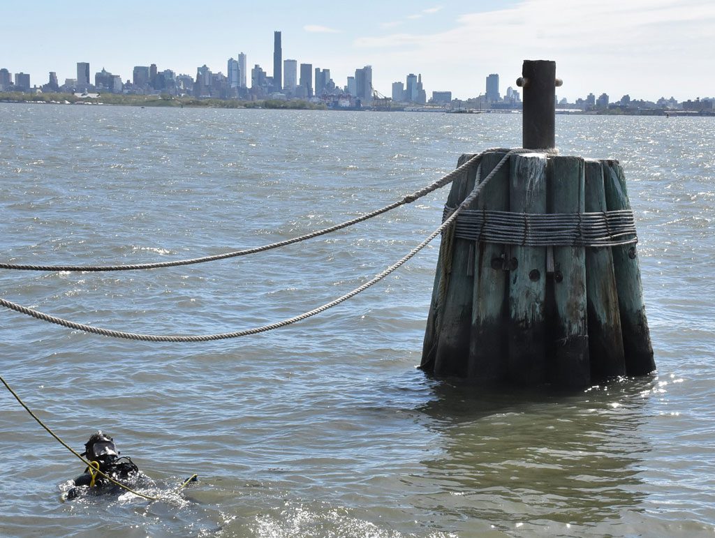 A U.S. Army diver from the 86th Engineer Dive Detachment prepares to inspect a dolphin at right (a cluster of piles driven into the ground serving as a fixed point to secure a vessel.)
