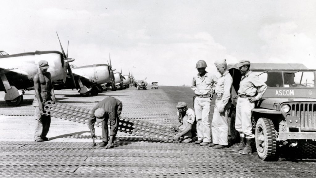 Construction of the critical Tanauan Airdrome, Leyte, Philippines, 16 December 1944. Men of the 1897th Engineer Aviation Battalion lay steel mat while commanding officer Col. James E. O'Keefe (left) and Maj. Gen. Hugh J. Casey (center) watch.