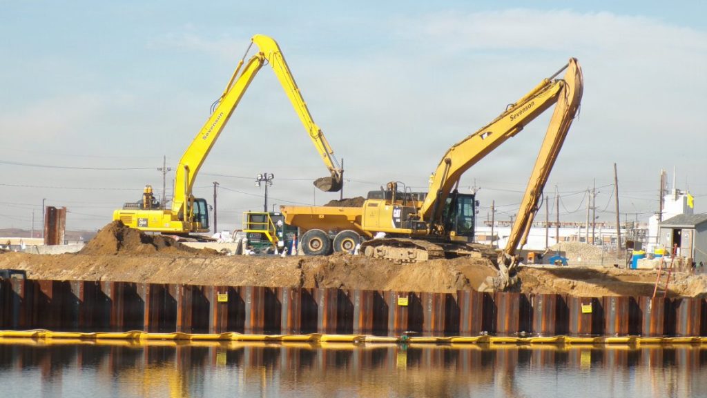 Two yellow long-reach excavators by Sevenson and Caterpillar moving soil into a dump truck at a remediation site behind a metal sheet pile wall along a waterfront.