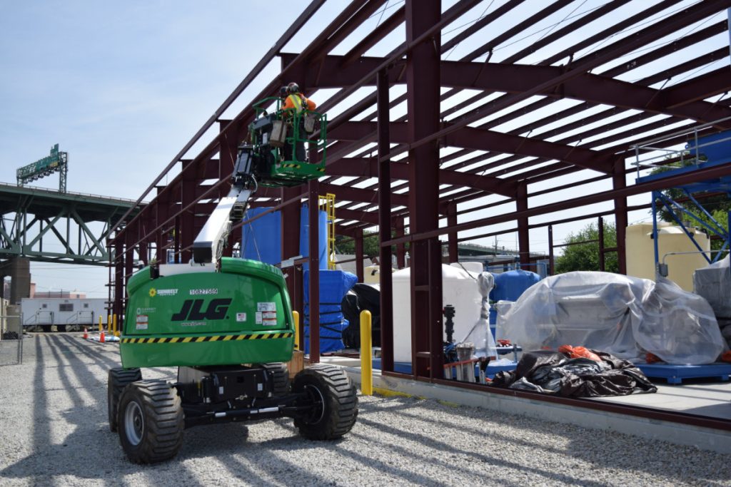 Two contractors in safety gear stand in the elevated basket of a green JLG boom lift, inspecting the steel frame of a large industrial building at a water treatment facility.