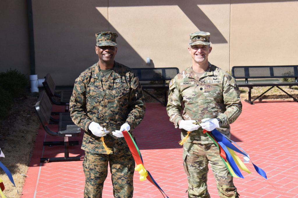 Two smiling military officers, one in a U.S. Marine Corps camouflage uniform and one in a U.S. Army OCP uniform, wearing white gloves and holding cut ceremonial ribbons on an outdoor red brick patio.