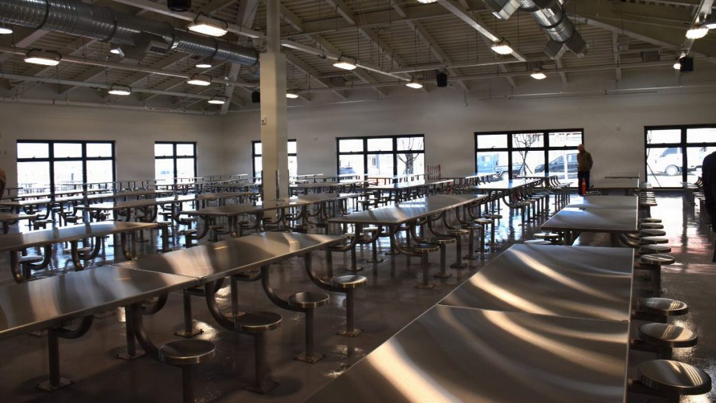 Wide interior view of an expansive, empty modern dining hall with rows of stainless steel tables, attached stool seating, high ceilings with exposed ductwork, fluorescent lighting, and bright large windows.