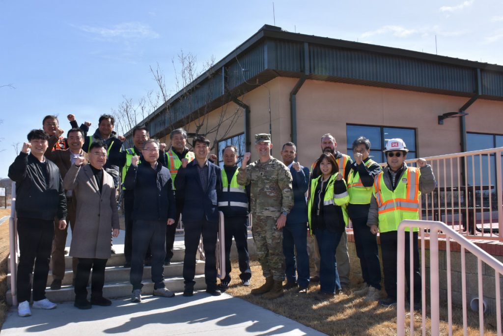 A group of approximately 15 people, including U.S. Army personnel in OCP uniforms and civilians in safety vests and business casual attire, standing on concrete steps in front of a modern tan building, smiling and raising their fists in a celebratory gesture.
