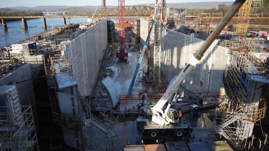 High-angle view looking down into a massive concrete lock chamber under construction with a large white Liebherr mobile crane positioned in the center and colorful shipping containers in the foreground.