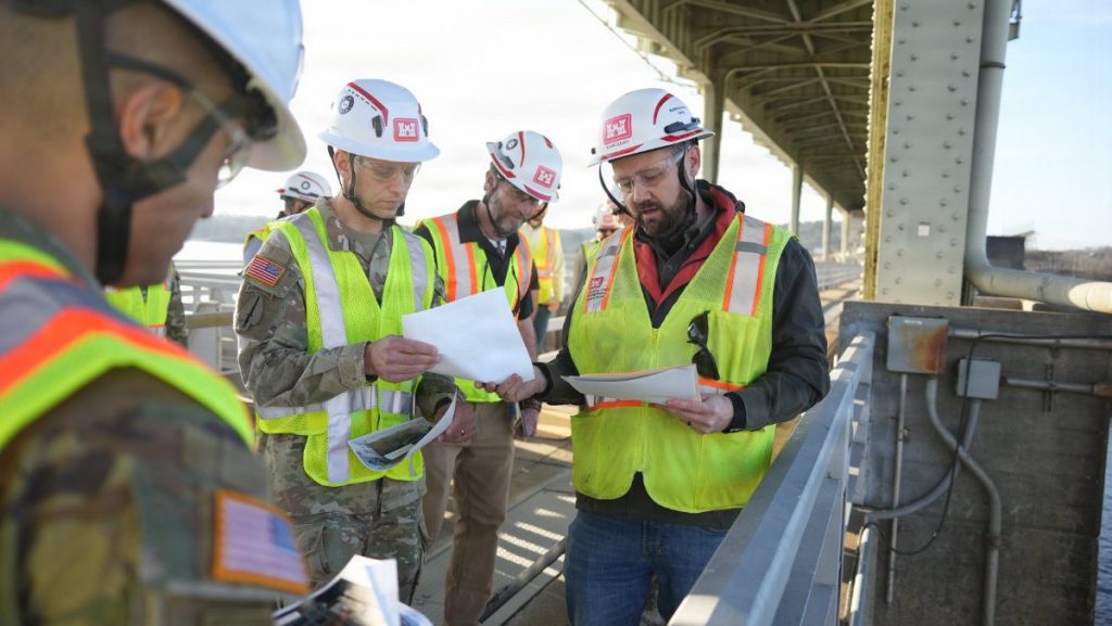Four men wearing USACE hard hats and high-visibility vests stand on an elevated walkway, huddled together while looking at and discussing printed technical documents.