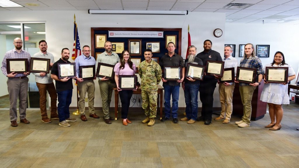 A diverse group of 14 people, including one U.S. Army officer in an OCP uniform and 13 civilians, standing in an office lobby while holding framed certificates in front of a Nashville District awards wall.