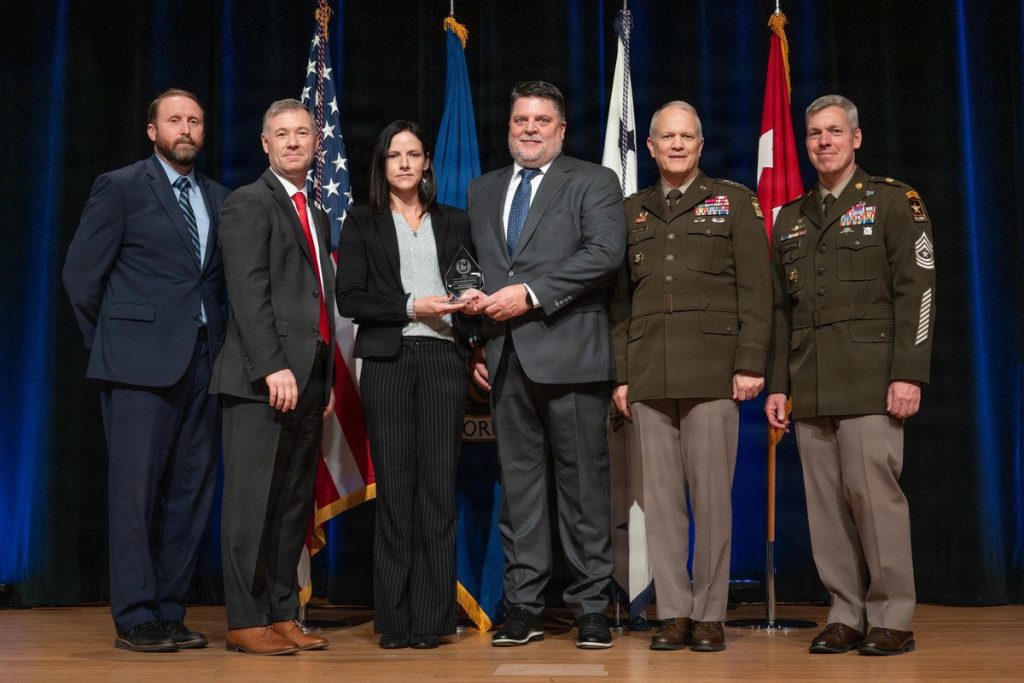 A woman and a man in professional attire holding a crystal award trophy on a stage, flanked by two military officers and two civilians.