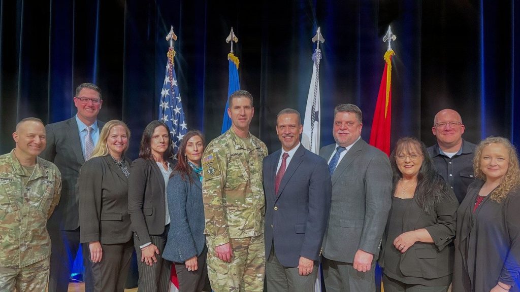 A group of eleven people, including two U.S. Army officers in OCP uniforms and nine civilians in professional attire, posing together on a stage in front of four flags and a dark blue curtain.