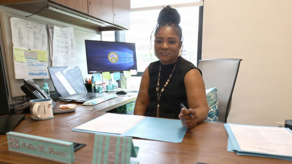 Senior HR Specialist Nakia Harris smiles while sitting at her office desk with folders and computers in the background.