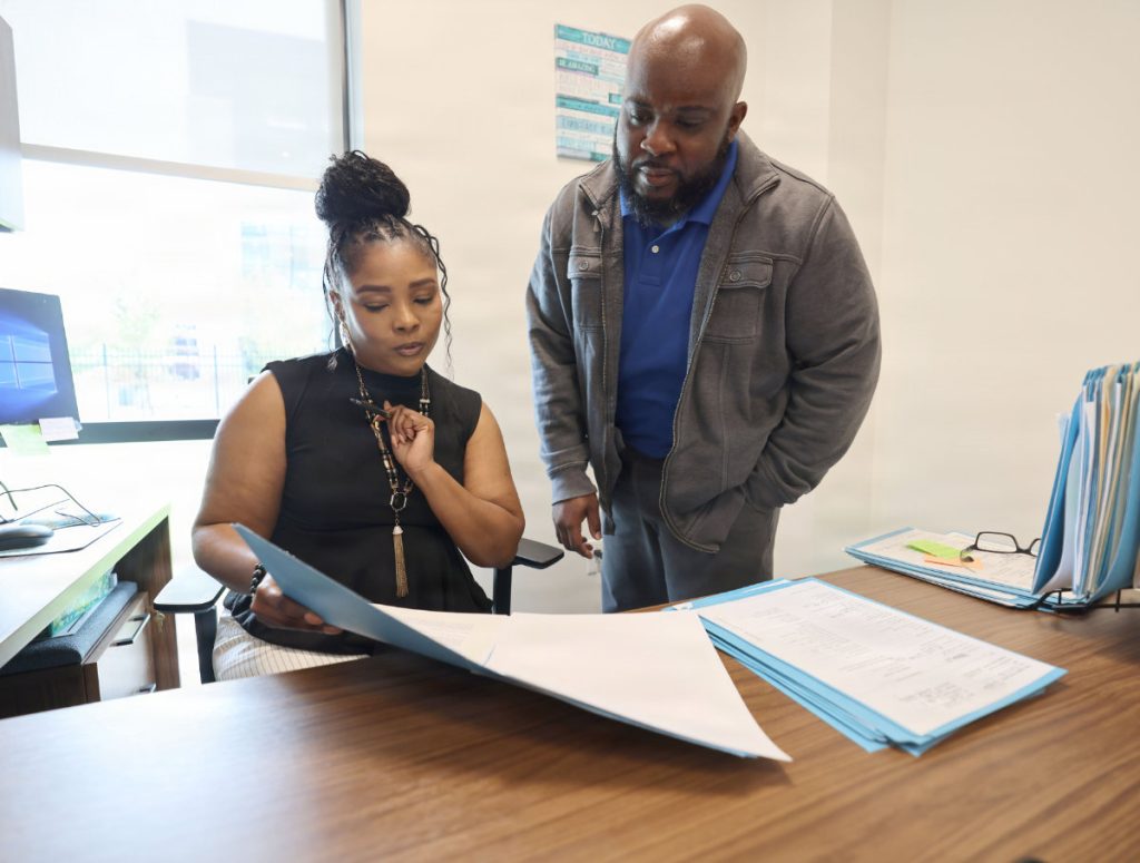 Nakia Harris stands beside a seated colleague in a human resources office while they both point to and review text on a printed sheet of paper.