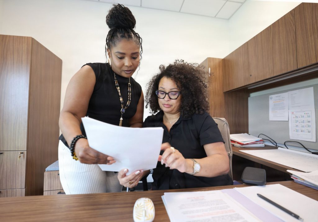 Senior HR Specialist Nakia Harris sits at her desk reviewing a folder with a colleague who stands beside her looking over the text in a bright office environment.