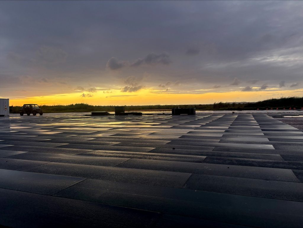 A vast grid of interlocking synthetic airfield matting reflecting the golden light of a dramatic sunset under dark, cloudy skies.