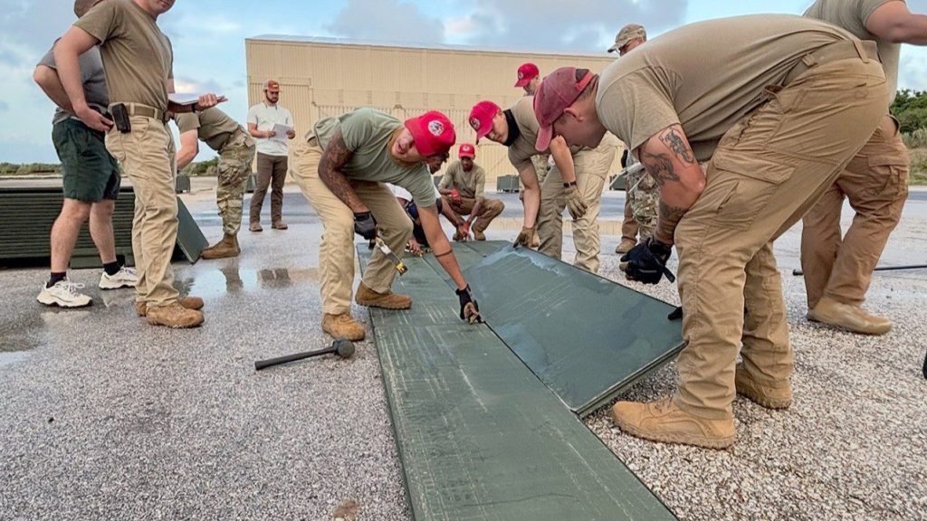 A team of military personnel in tan uniforms and red hats work together to assemble and lock green interlocking airfield mats on a concrete surface.