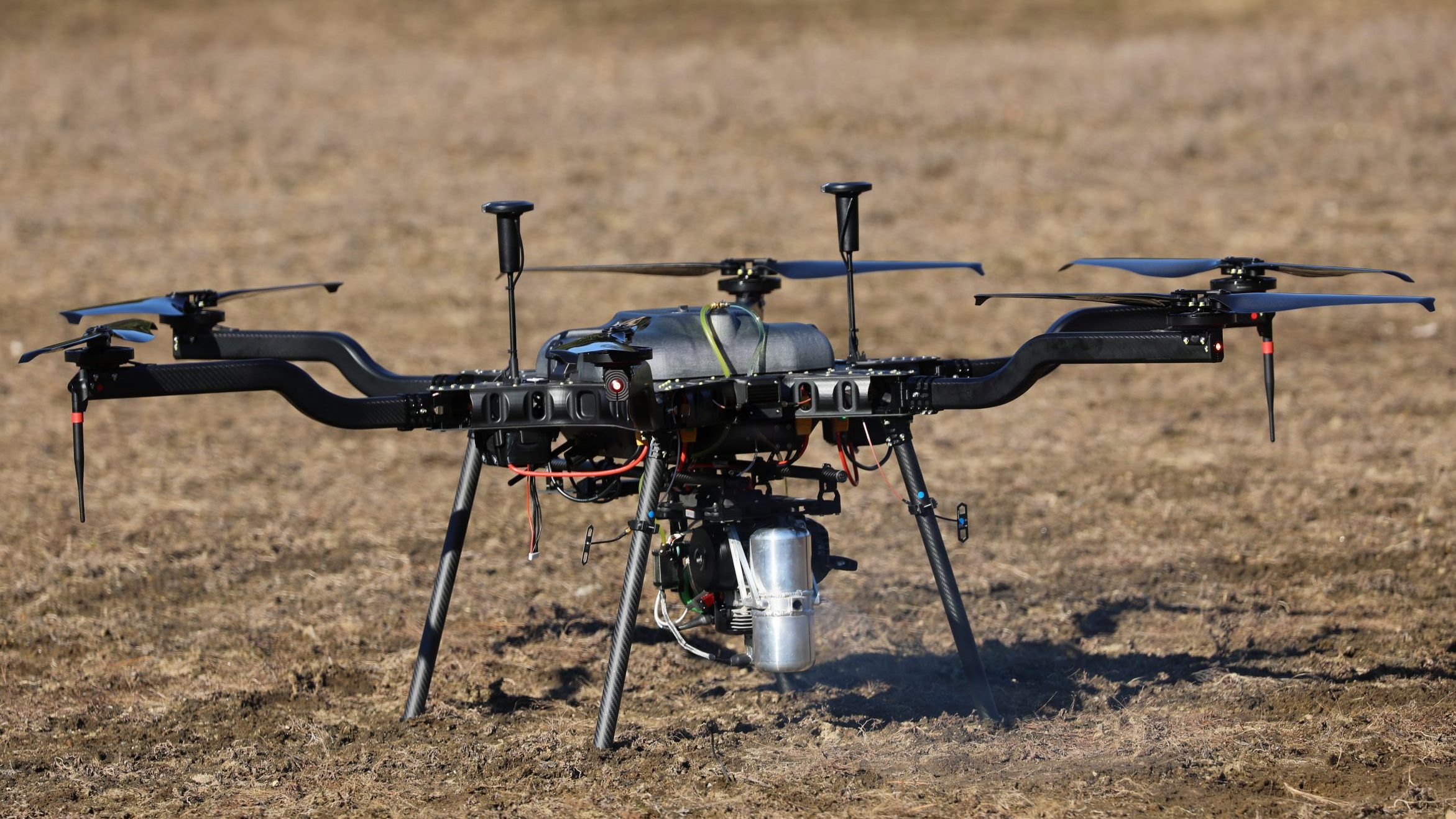 A detailed close-up of a heavy-lift hexacopter drone resting on its carbon-fiber landing gear on dry grass, showcasing its hybrid gas-electric engine setup.
