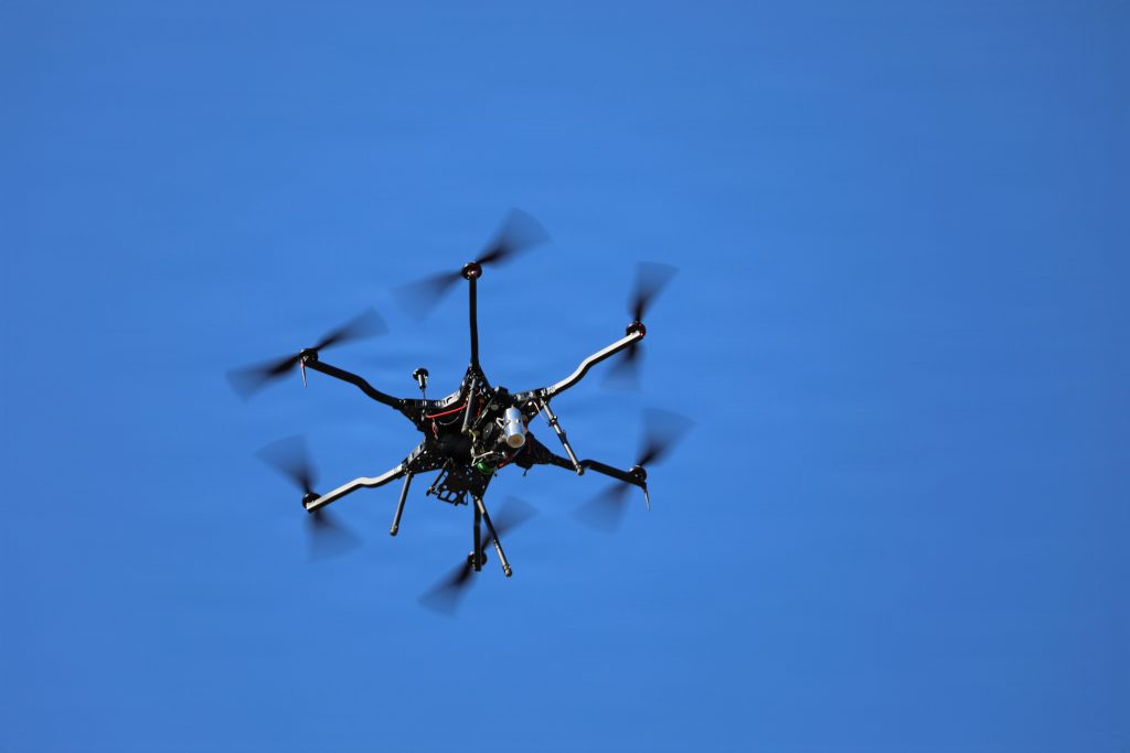 An under-vessel view of a large black multi-rotor hexacopter drone flying mid-air with spinning propellers against a solid, clear blue sky.
