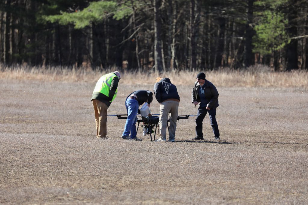 Four personnel, including one wearing a high-visibility safety vest, stand in an open winter field bending over a large multicopter drone to perform field calibration.