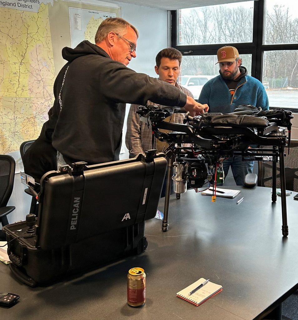 Three men gather around a large, black industrial hexacopter drone resting on a conference table next to an open rugged Pelican equipment case.