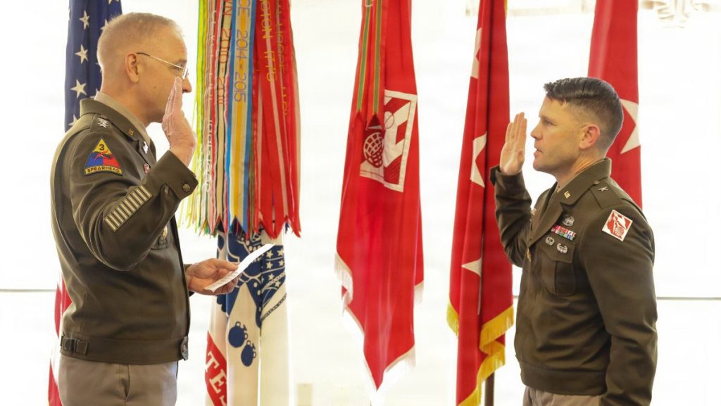 Two U.S. Army officers in dress uniforms face each other with their right hands raised to administer an official military oath, flanked by ceremonial organizational flags.