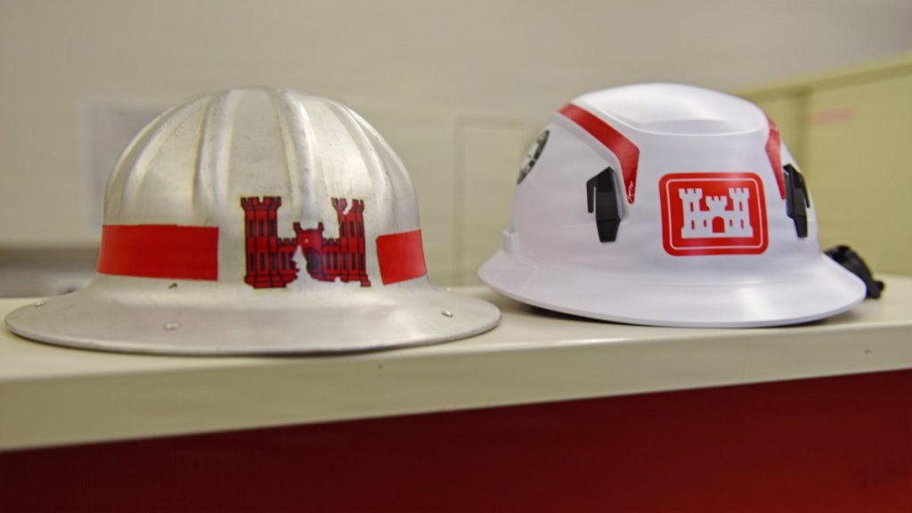 A vintage metal USACE hard hat from the 1930s-1950s sits next to a modern white U.S. Army Corps of Engineers safety helmet, both featuring the red castle logo.