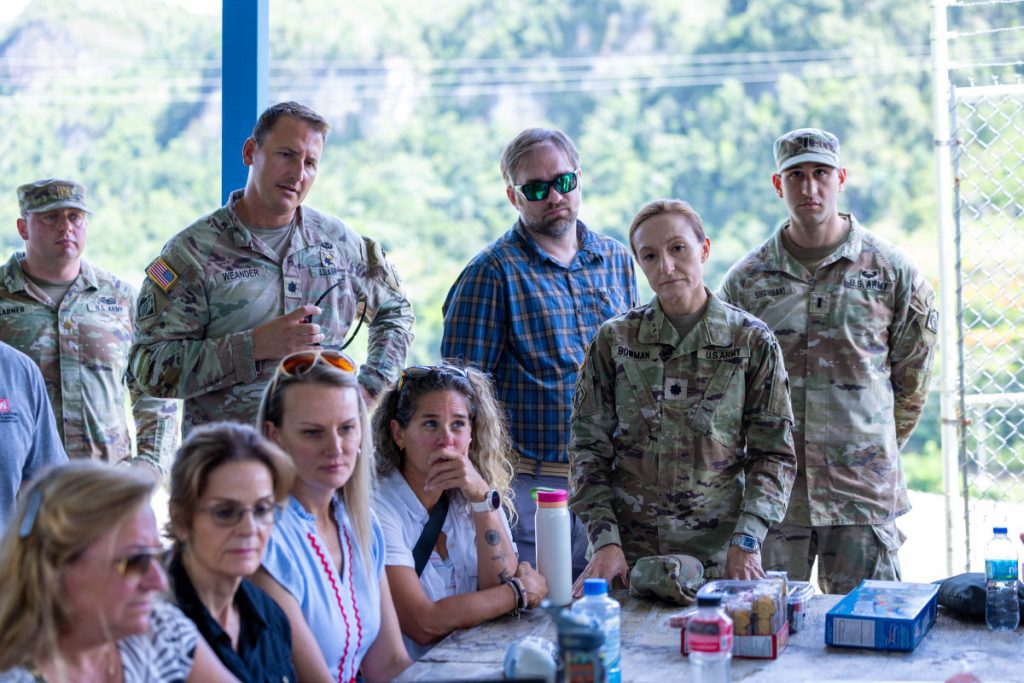 A group of military personnel in camouflage uniforms and civilians sit outdoors under a canopy structure, listening attentively during a briefing.