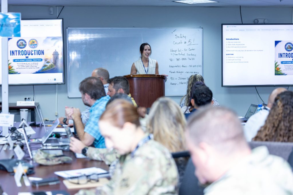 A woman stands at a wooden podium speaking to a classroom style setup of military personnel and civilians working on laptops at long conference tables.