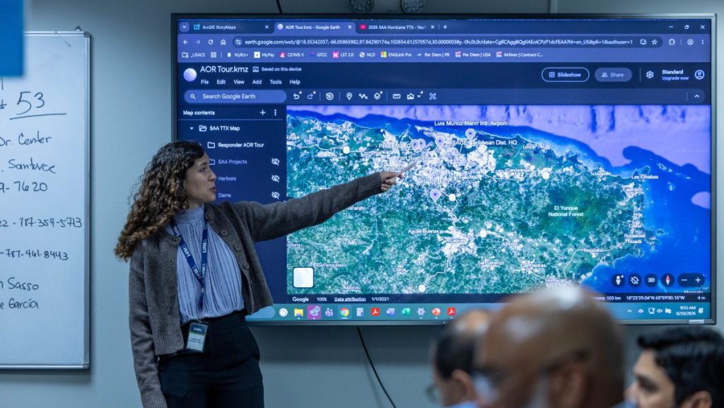 A woman stands in front of a large display monitor, pointing out geographical locations on a digital satellite terrain map of Puerto Rico for an audience.