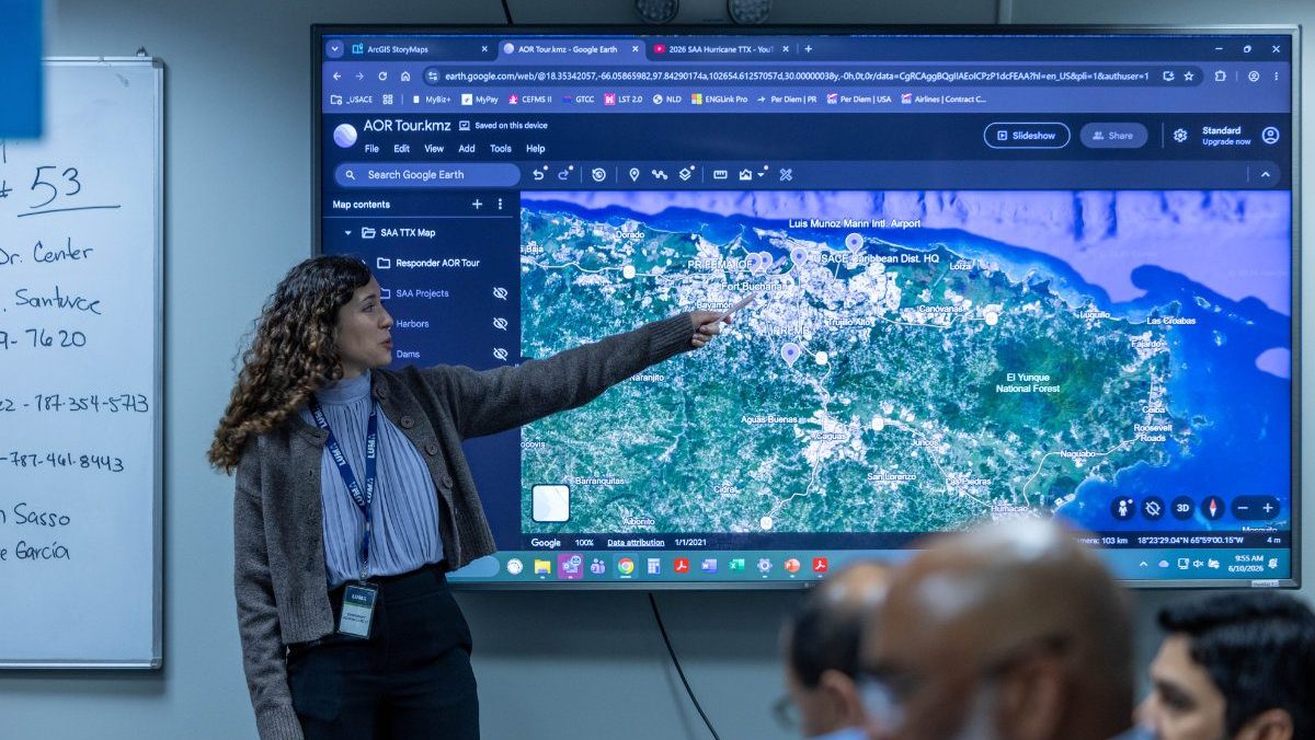 A woman stands in front of a large display monitor, pointing out geographical locations on a digital satellite terrain map of Puerto Rico for an audience.