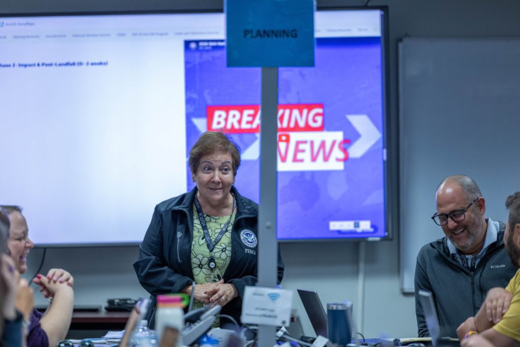 A woman wearing a FEMA identification lanyard speaks to a group gathered around a conference table, with a "Breaking News" graphic visible on a screen behind her.