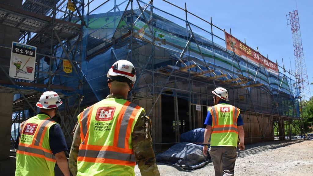 Three individuals wearing white hard hats and high-visibility vests with the US Army Corps of Engineers Far East District logo walk toward a multi-story building covered in construction scaffolding and blue safety netting.