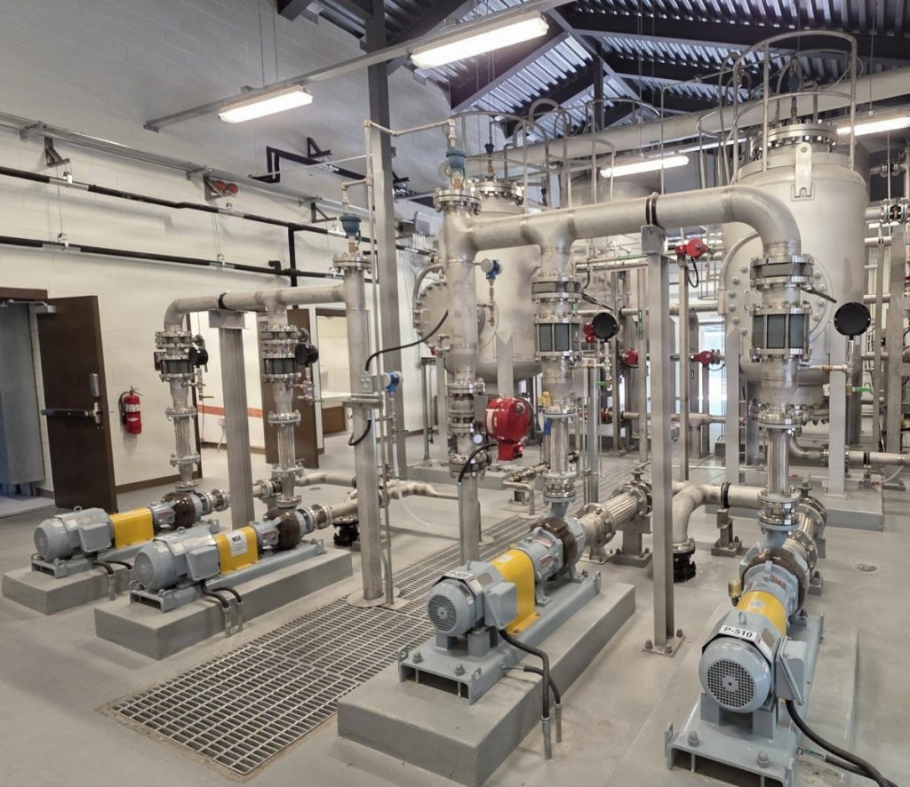 The interior of an industrial facility featuring rows of grey and yellow electric water pumps mounted on concrete bases, connected to a complex network of stainless steel piping, valves, and large filtration tanks.