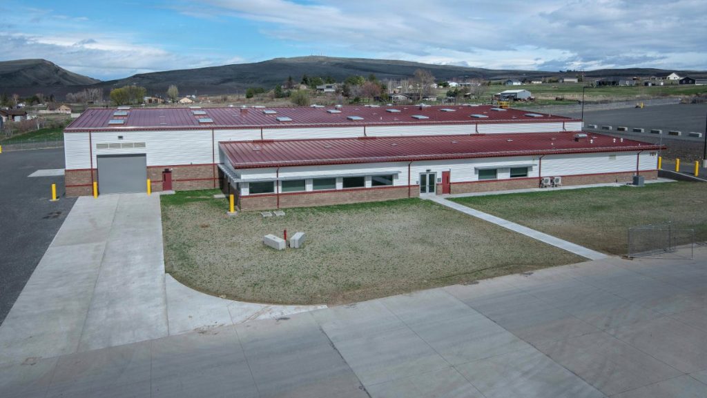 High-angle exterior view of a modern industrial military facility building featuring a white corrugated facade, a red metal roof, and a clean concrete driveway.