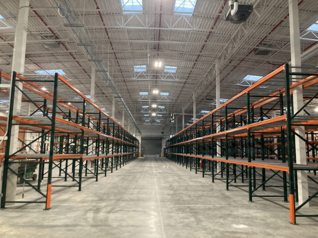 Interior of a vast, empty industrial warehouse featuring high ceilings with skylights and long rows of dark green and bright orange storage racks.