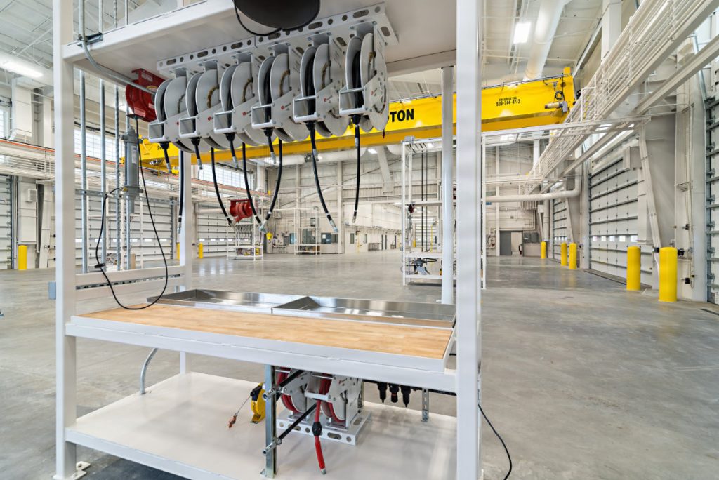 A modern vehicle maintenance shop interior showing a central wooden workbench with overhead fluid hose reels and a large yellow 10-ton overhead crane.