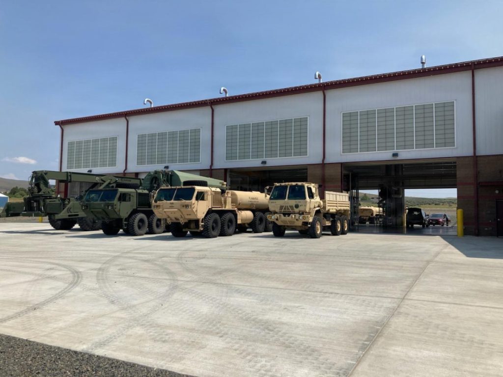 Several large military tactical vehicles, including tan supply trucks and a fuel tanker, parked on a concrete apron outside the open bay doors of a maintenance facility.