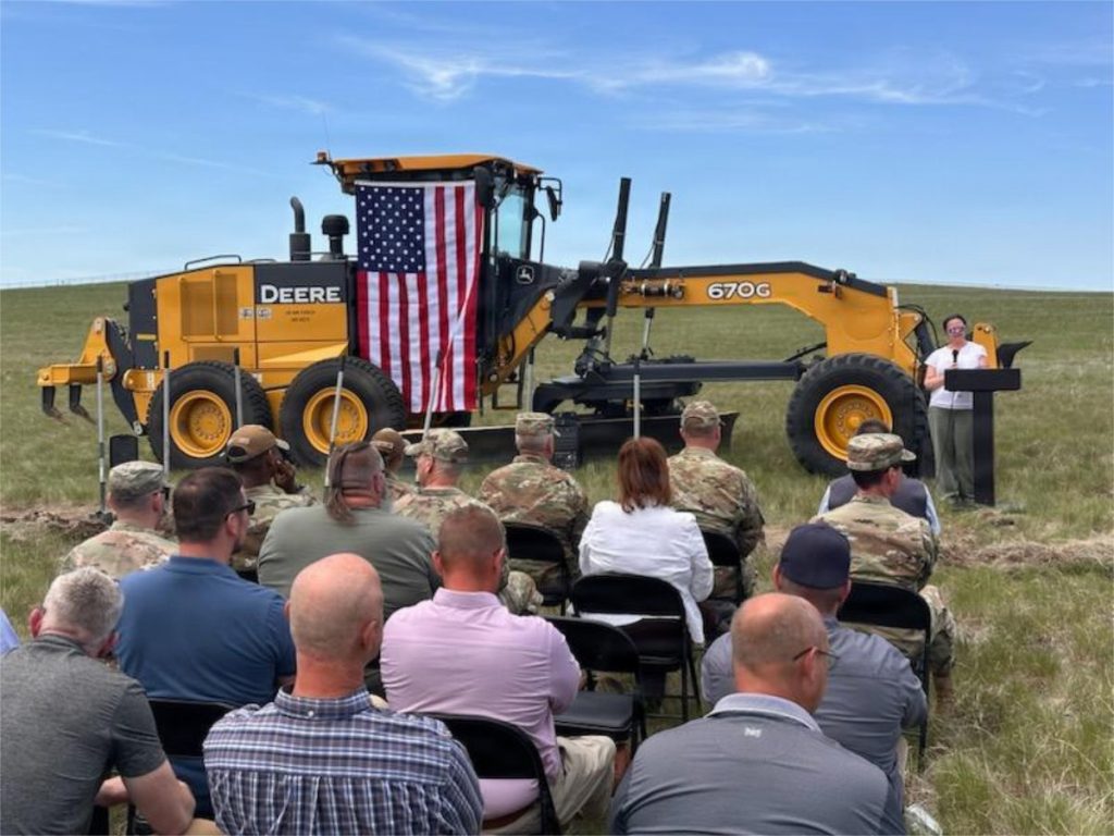 An audience of military personnel and civilians sits in folding chairs outdoors, watching a woman speak at a podium next to a large yellow John Deere tractor draped with an American flag.