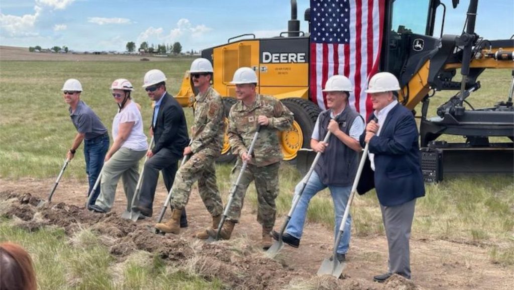 Seven individuals in hard hats, including military officers and civilians, stand in a line using shovels to break ground in a grassy field next to a large yellow tractor.