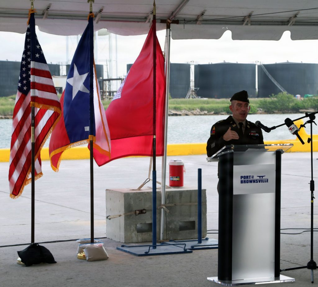 A U.S. Army officer in dress uniform speaks at a podium branded with the Port of Brownsville logo, standing next to the American flag, Texas state flag, and a red USACE flag under an outdoor tent.