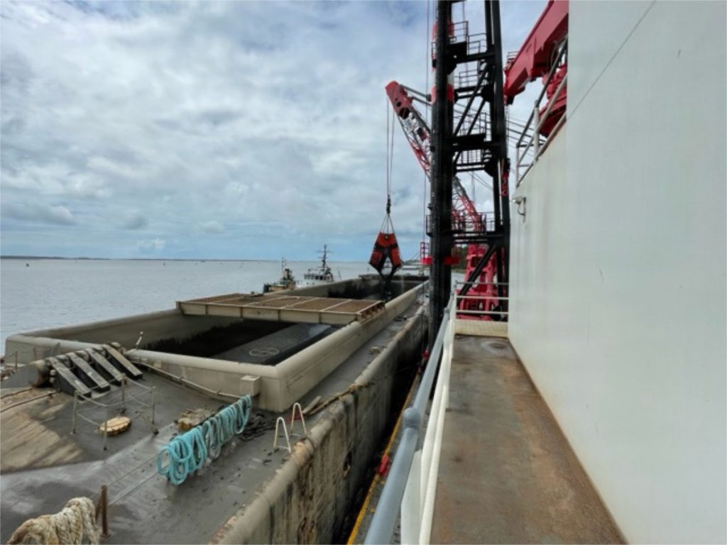 Looking down from the deck of a larger ship alongside an industrial hopper barge, where a heavy mechanical clamshell bucket crane hovers over the open cargo hold.