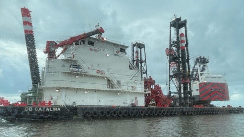 A large industrial dredging vessel named DB CATALINA floats on choppy water under an overcast sky, equipped with heavy cranes, rigging towers, and red-and-white striped safety markers.