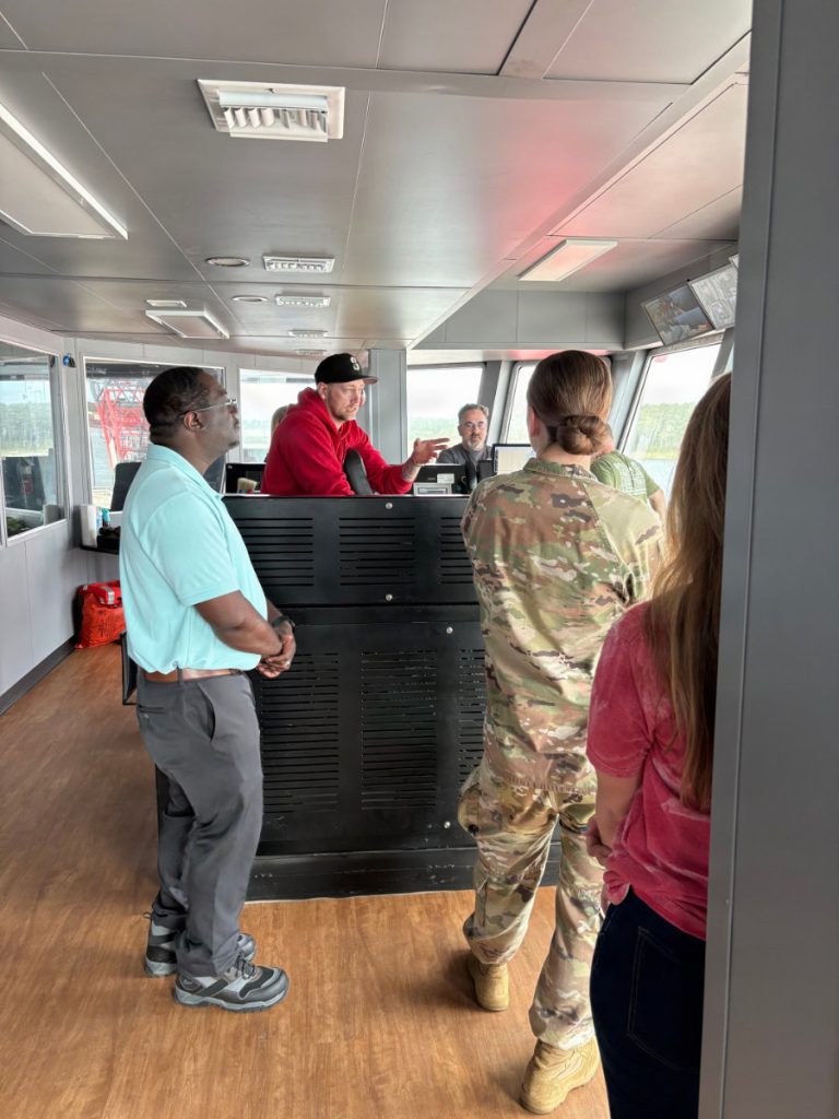 Inside the bridge of a vessel, a man in a red hoodie gestures while speaking to visitors, including a person in an Army camouflage uniform and a civilian in a light blue polo shirt.