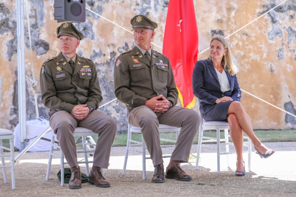Two U.S. Army officers in formal dress uniforms and a woman in a dark business suit sit in white chairs on an outdoor stage during a military ceremony.
