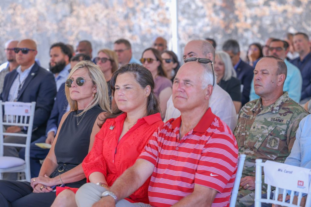 An audience of military personnel and civilians sit in white chairs, watching an outdoor official ceremony with attention.