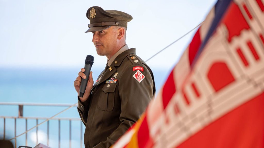 An Army officer in a service uniform stands at a podium, holding a microphone and speaking at an outdoor ceremony, with a blurred USACE flag in the foreground.