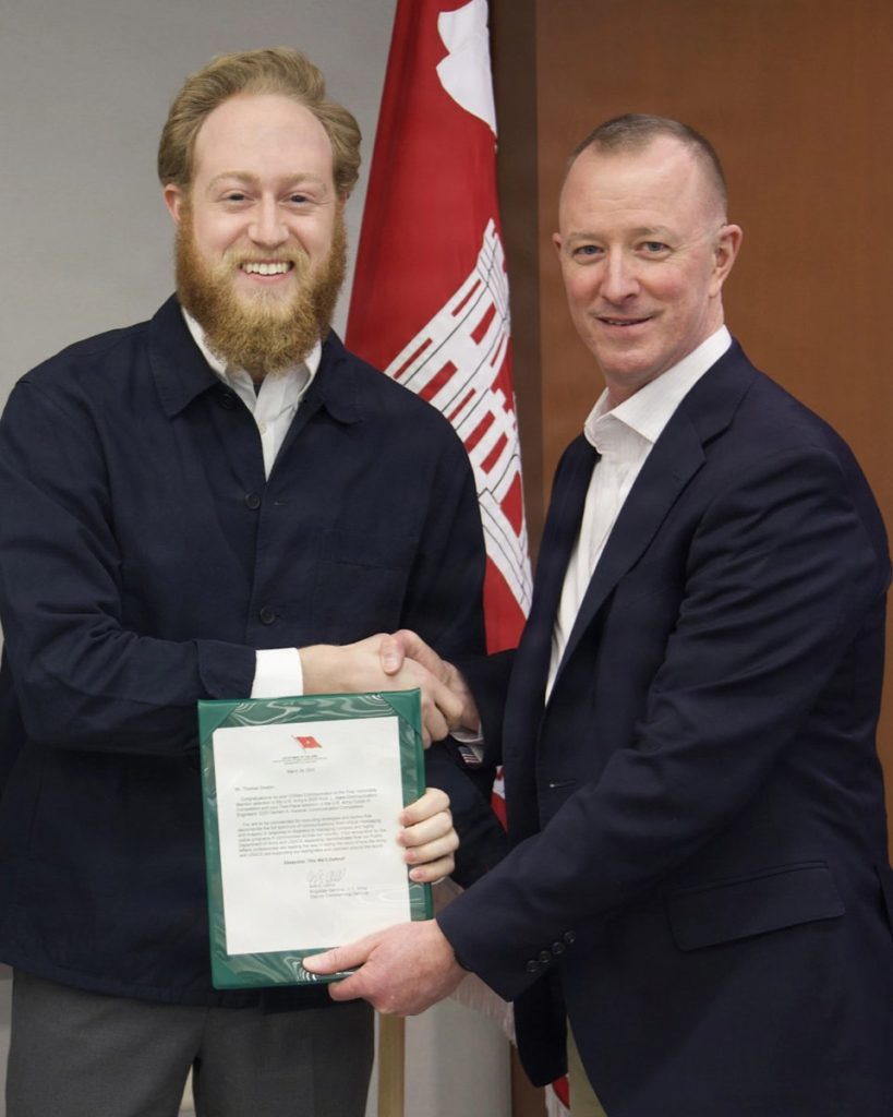 A bearded public affairs specialist smiles and shakes hands with an official in a navy suit jacket while presenting a framed certificate of achievement in front of a red USACE flag.