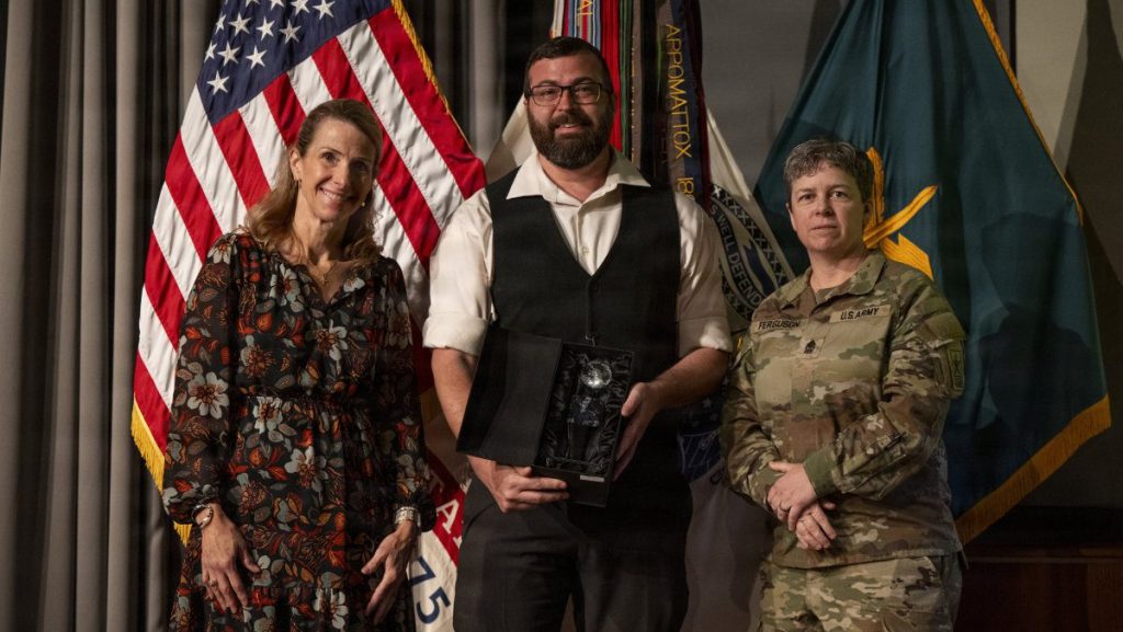 A award recipient wearing glasses and a dark vest holds a boxed crystal trophy while standing between a civilian woman in a patterned dress and a U.S. Army senior officer in camouflage uniform in front of military flags.