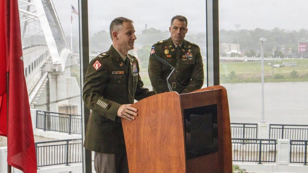 An Army officer stands at a wooden podium speaking into a microphone, while another officer stands behind him, in front of a large window overlooking a bridge and a river.