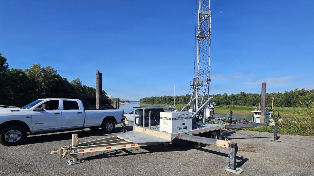 A mobile trailer platform with extended stabilizer legs and an attached communications mast parked near a white pickup truck along a riverbank.