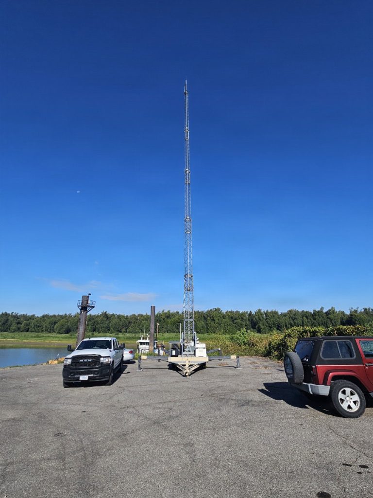 A tall lattice communications tower fully extended upward from a mobile trailer setup parked in a asphalt lot near a waterway.