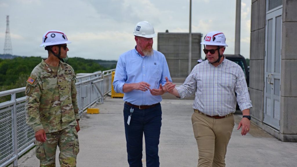 An Army officer in camouflage uniform, a TVA official in a blue shirt, and ASA Civil Works Adam Telle in a plaid shirt walk together across a dam pathway while wearing hard hats.