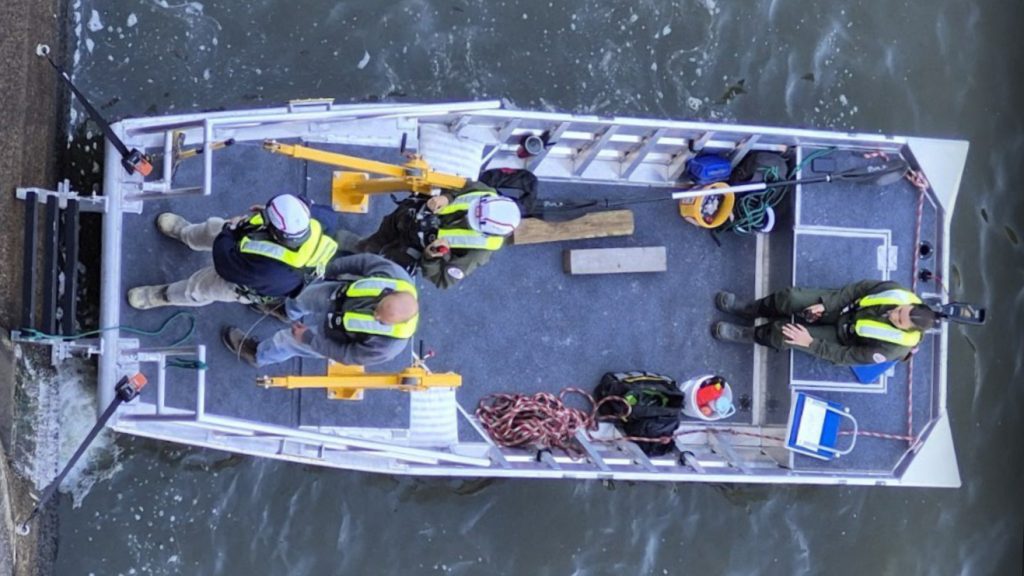 An overhead aerial view of four rescue team members wearing life jackets and helmets inside a small, flat-bottomed aluminum workboat moored against a concrete wall.